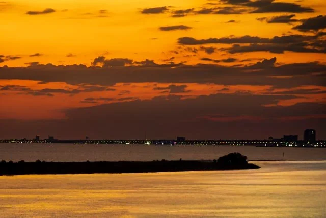 Golden-orange sunset over the Mississippi Gulf Coast with dark clouds, reflective water, and a distant shoreline silhouetted on the horizon.