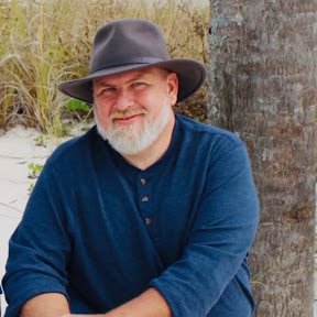 Portrait of Kenneth D. Hanks seated outdoors beside a tree, wearing a brown hat and blue shirt.