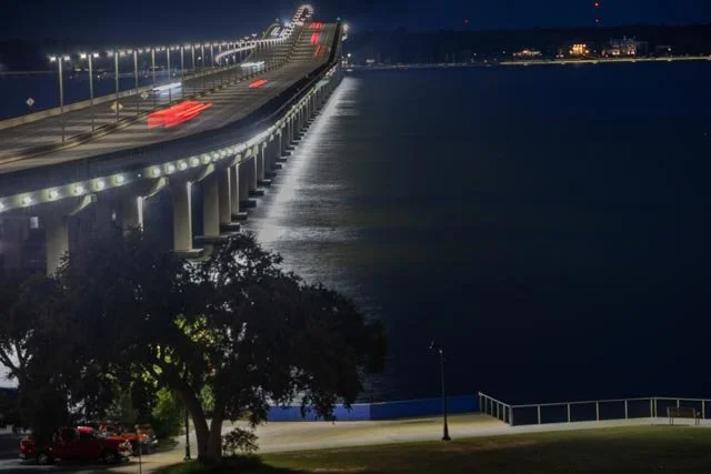 Night photograph of a brightly illuminated bridge stretching across dark water on the Mississippi Gulf Coast, with red vehicle light trails and reflections shimmering below.