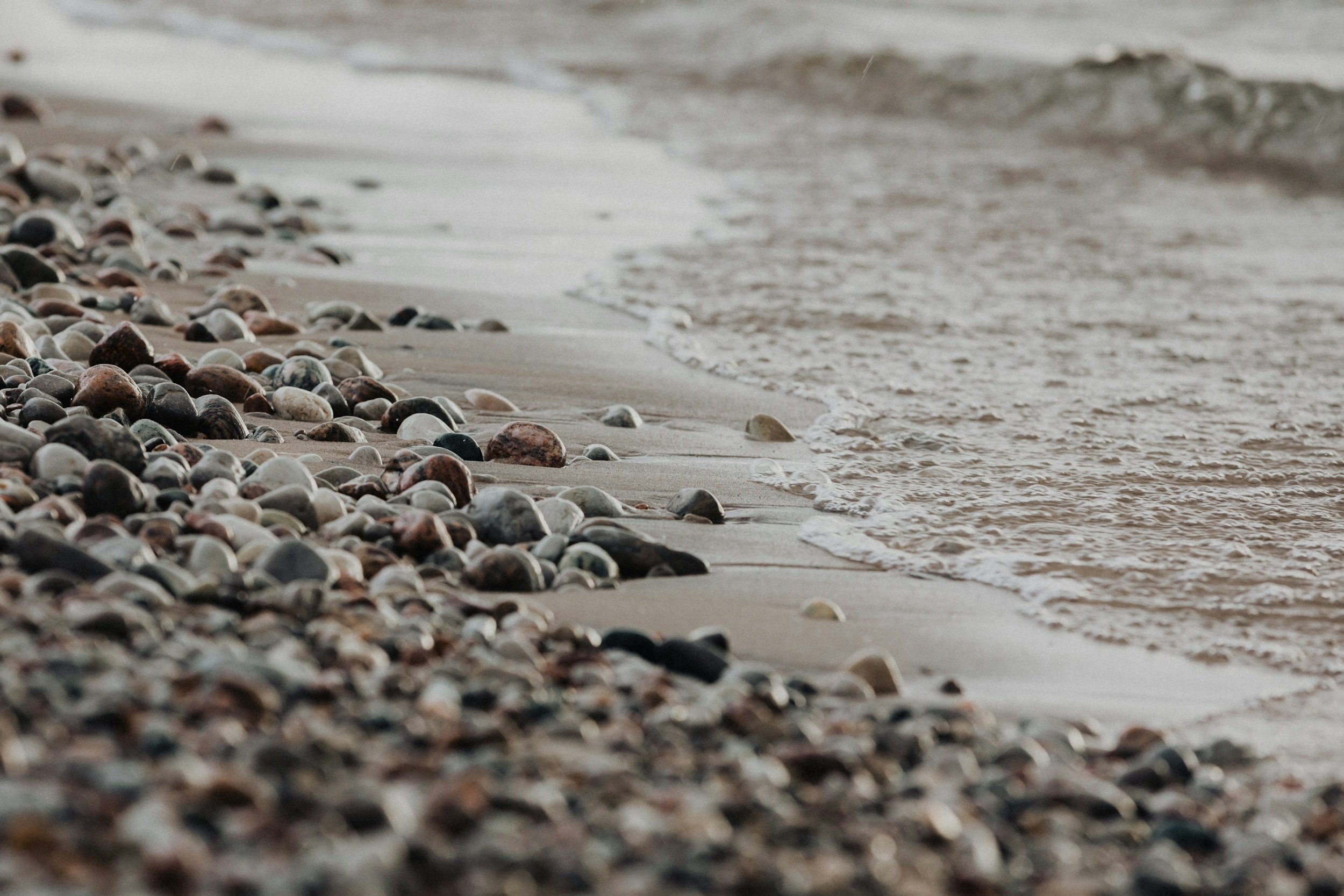 A close-up view of a rocky beach with multicolored stones, gentle waves, and sandy shore, with the ocean in the background.