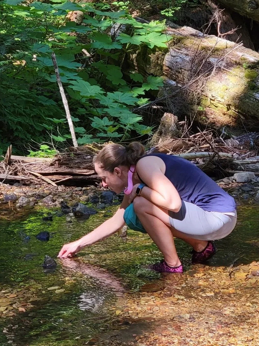 A woman crouches in a shallow creek, reaching into the water with her hand. She is wearing a navy tank top, beige shorts, and purple shoes, with a smartwatch on her wrist. The creek is surrounded by green foliage, rocks, and fallen tree branches.