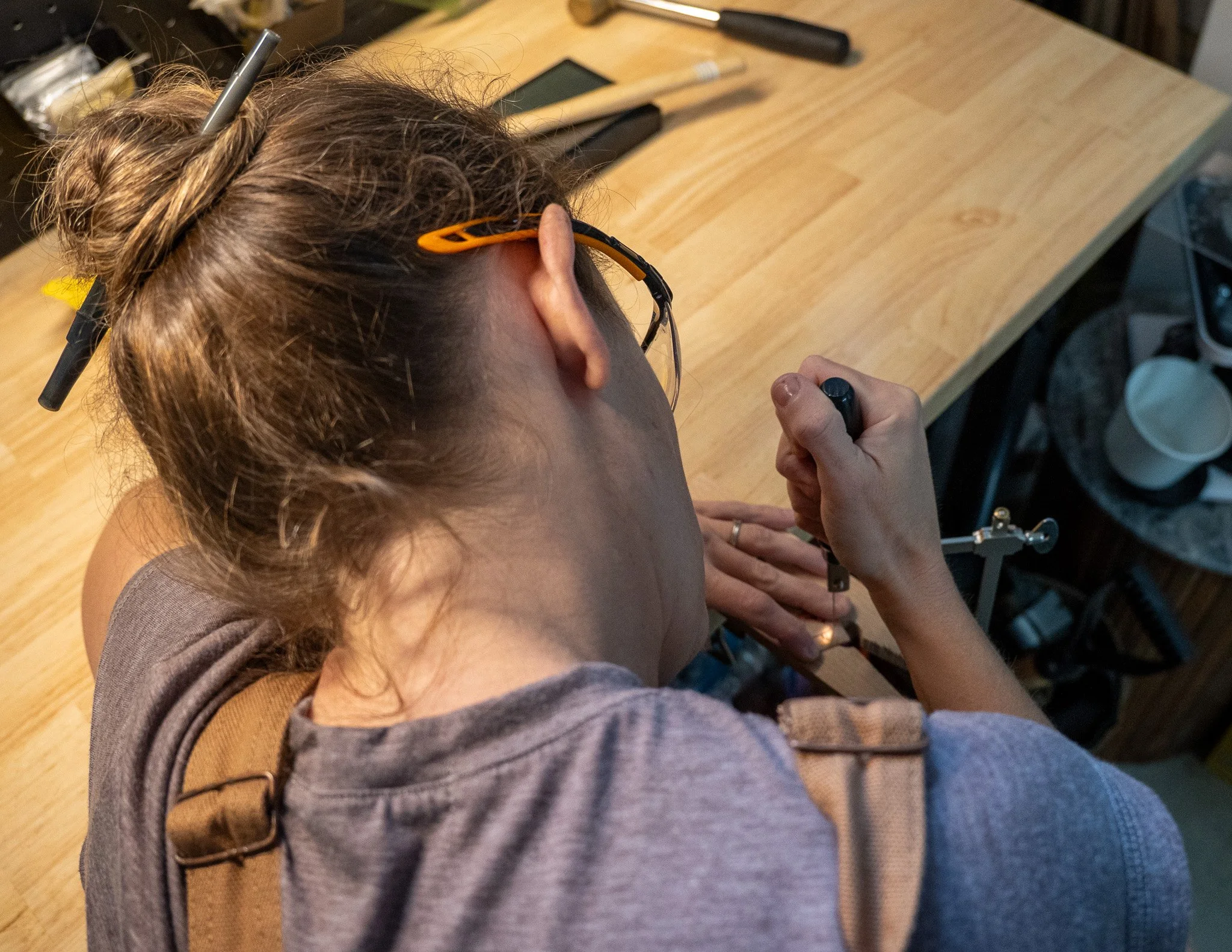 Top view of a woman with brown hair styled in a braid, wearing glasses with orange accents, working with a tool at a wooden workbench.