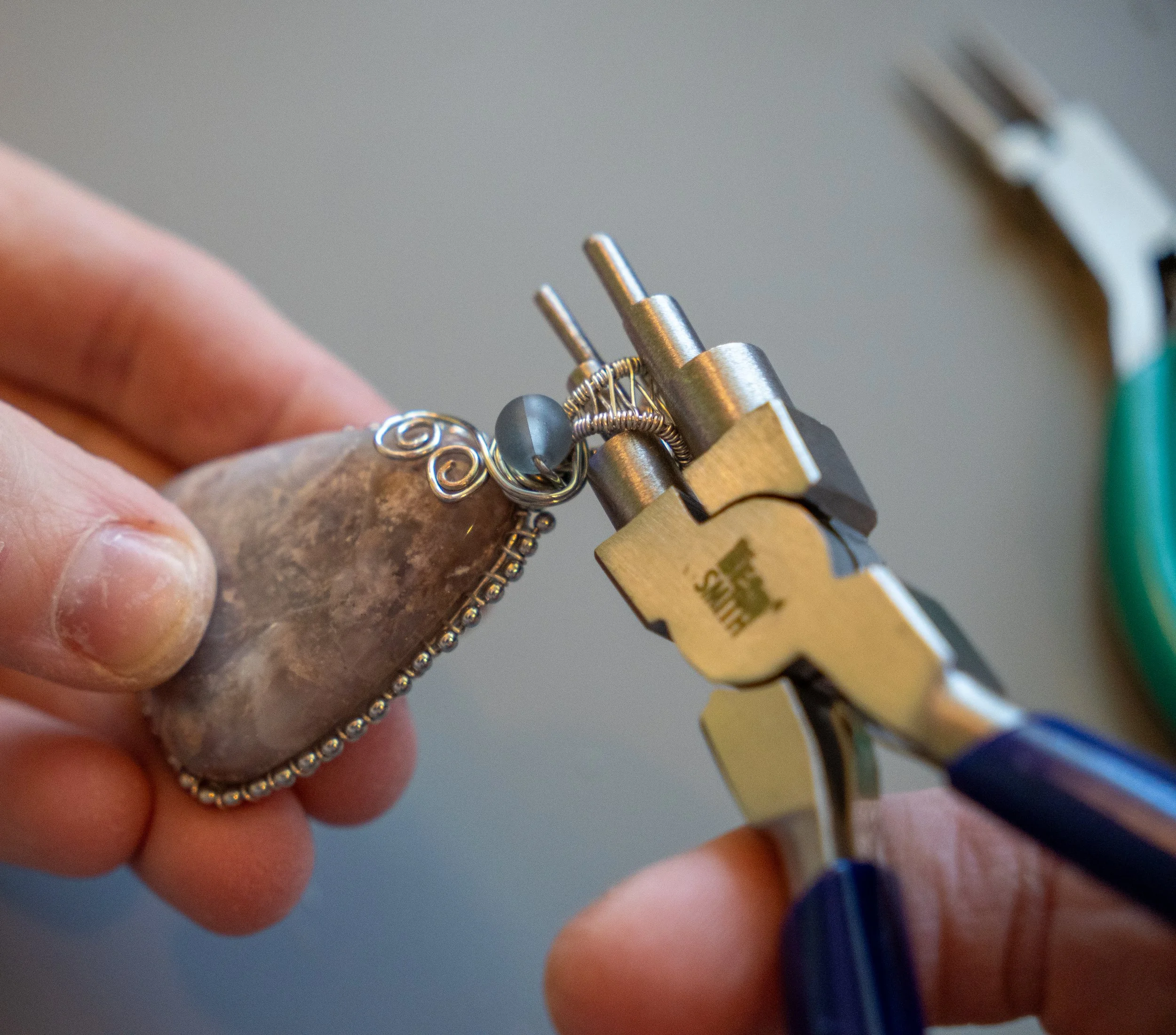 Close-up of someone's fingers holding a wire jewelry piece near a small clamp tool.