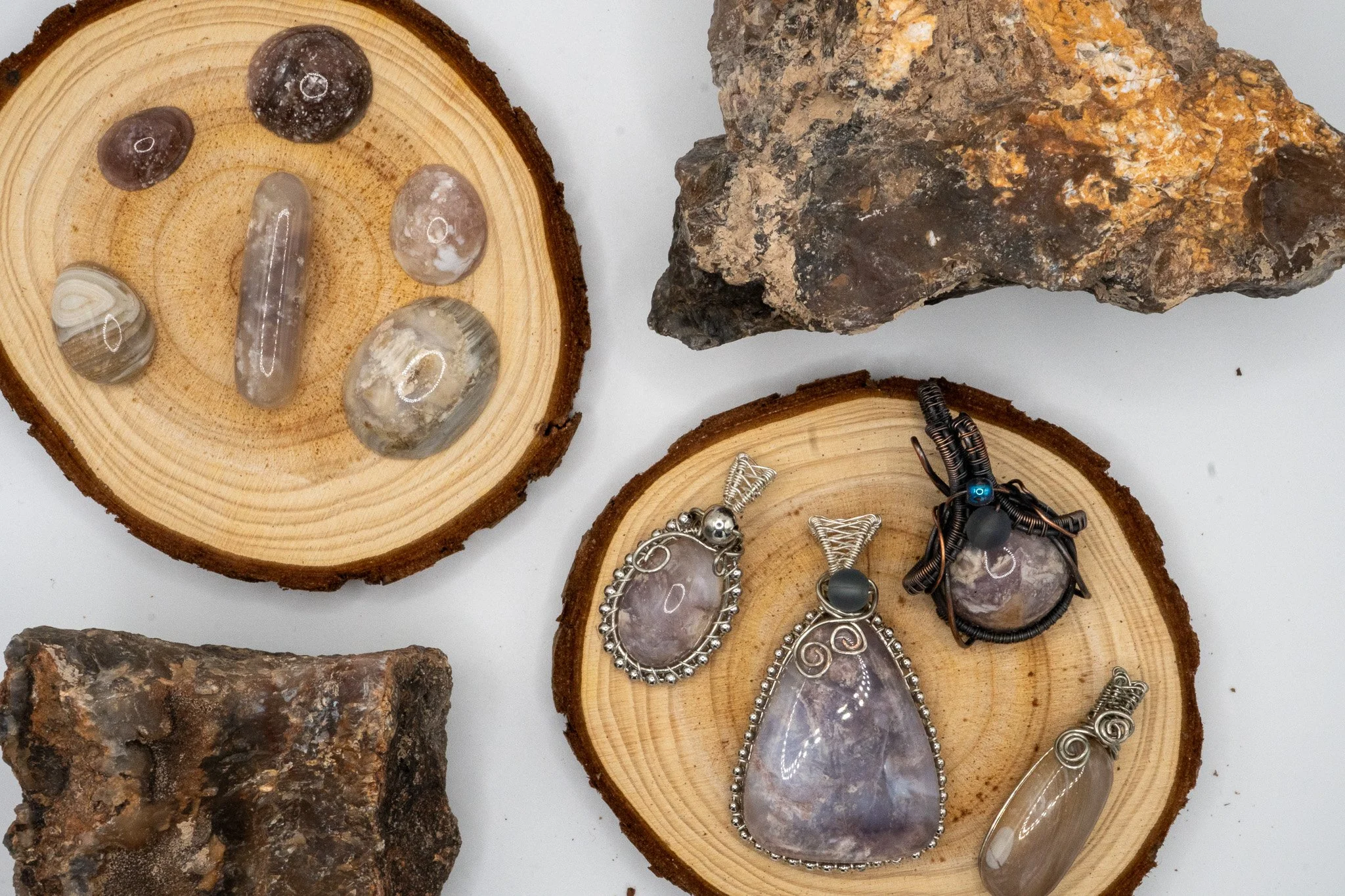 Several pieces of polished and raw stones displayed on wooden slices, with some jewelry including necklaces and rings made from wire and stones.
