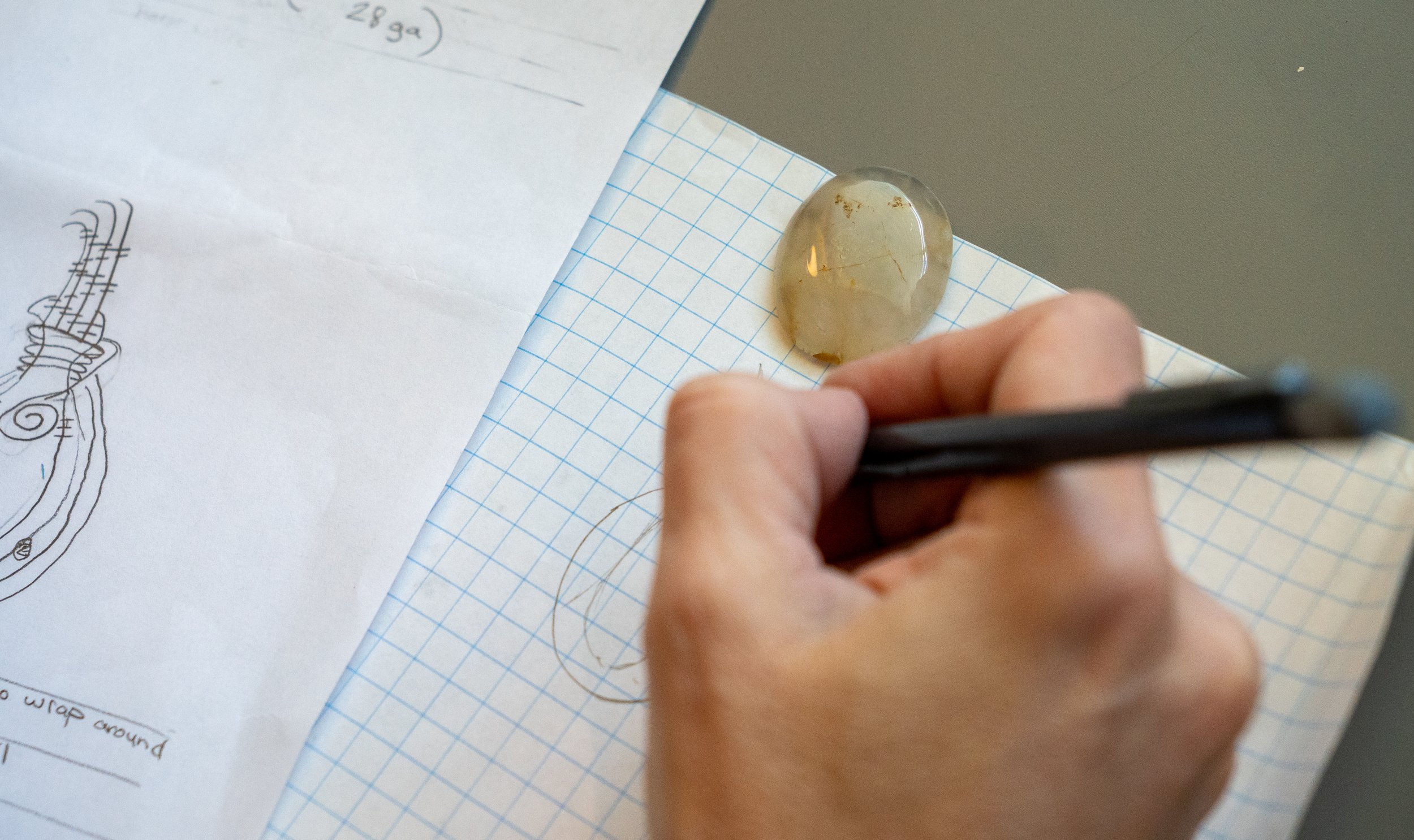 A person's hand holds a black pen and writes on a piece of graph paper with notes and sketches. Several sheets of paper, including a drawing and handwritten notes, are laid out on a flat surface. A glass paperweight with some discoloration or stains rests on the graph paper.