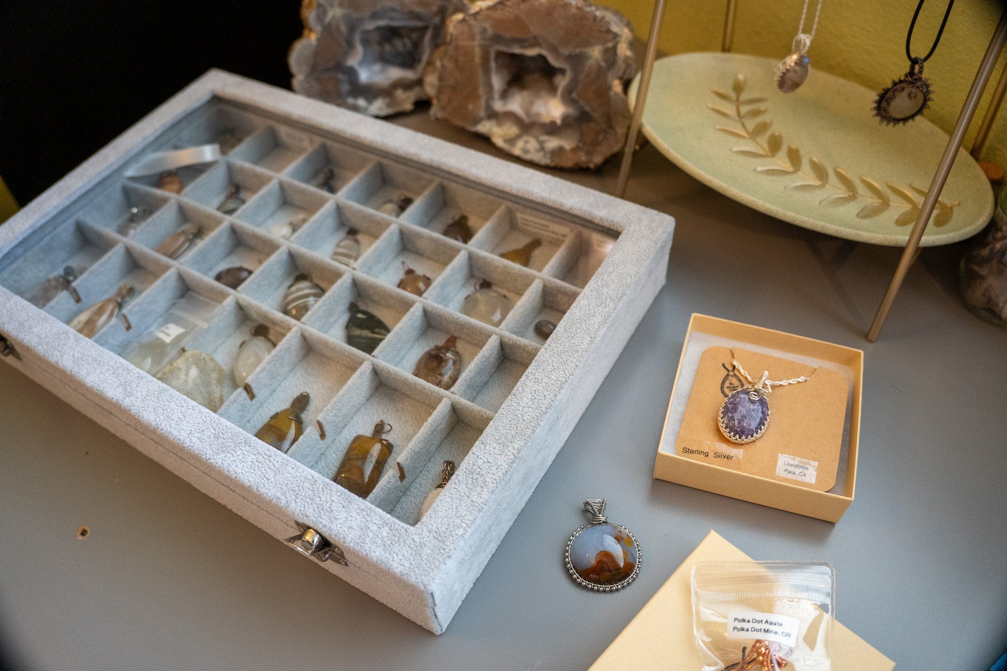 Display of various jewelry pieces, including a large amethyst pendant necklace, in glass cases and on display cards, with natural rock formations in the background.