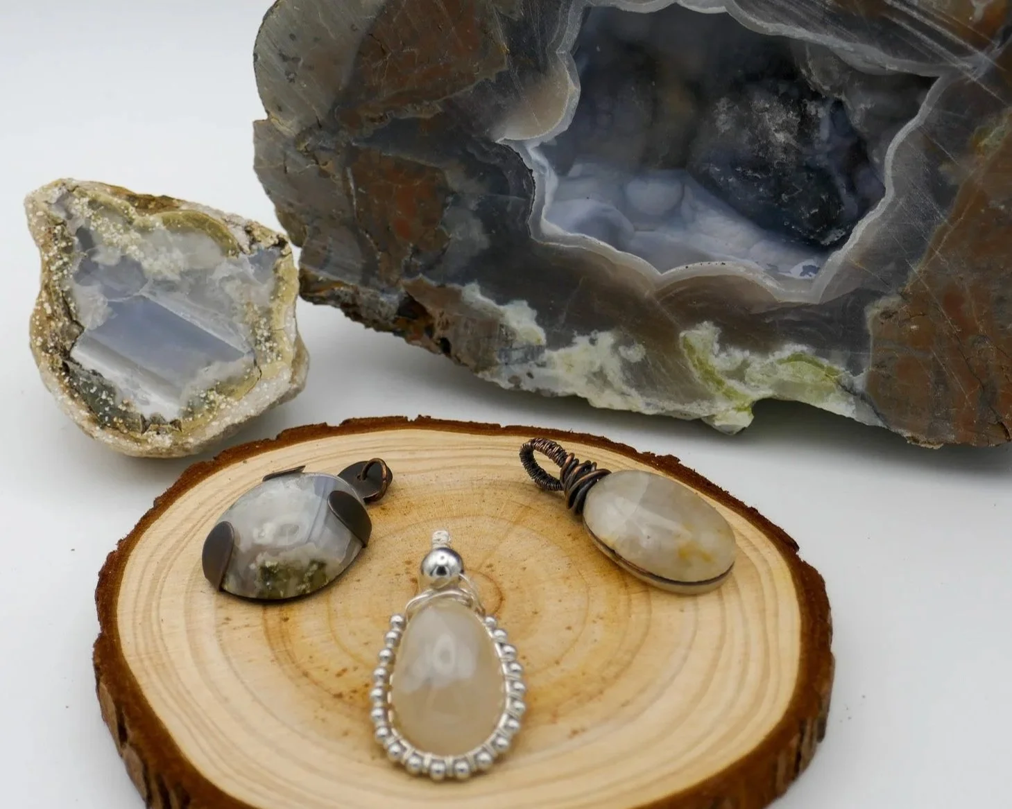 A collection of jewelry and geodes on a wooden surface with a white background. The jewelry includes a pendant with a large beige stone, a ring with a large stone, and a small pendant with a gray stone. Two geodes are visible, one cut open displaying crystals inside, and the other larger, rough, and uncut.