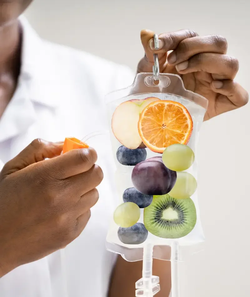 Hand holding clear bag with artificial slices of orange, apple, blueberry, green grape, purple grape, and kiwi, attached to a medical IV pole.