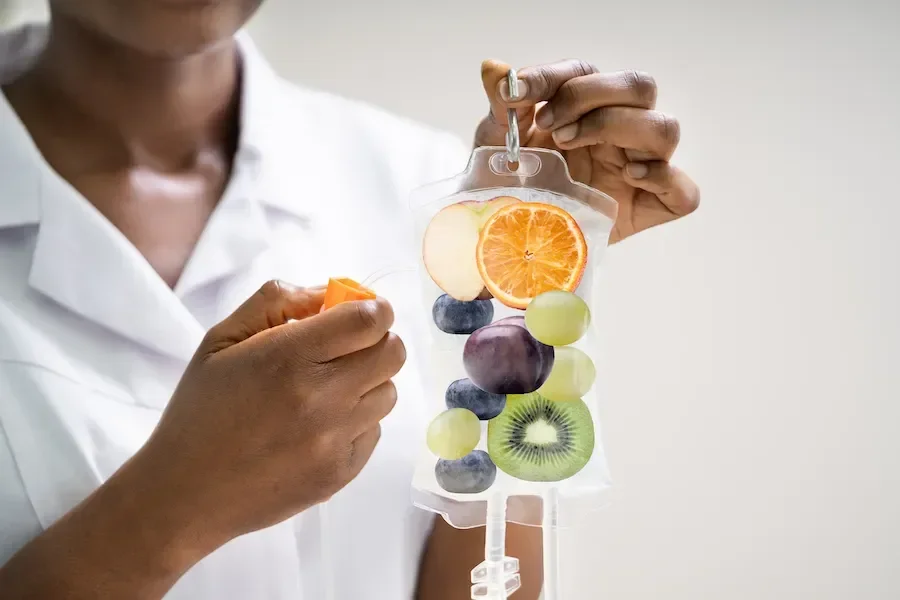 Person holding a transparent IV bag filled with slices of various fruits including orange, green grape, grape, and kiwi