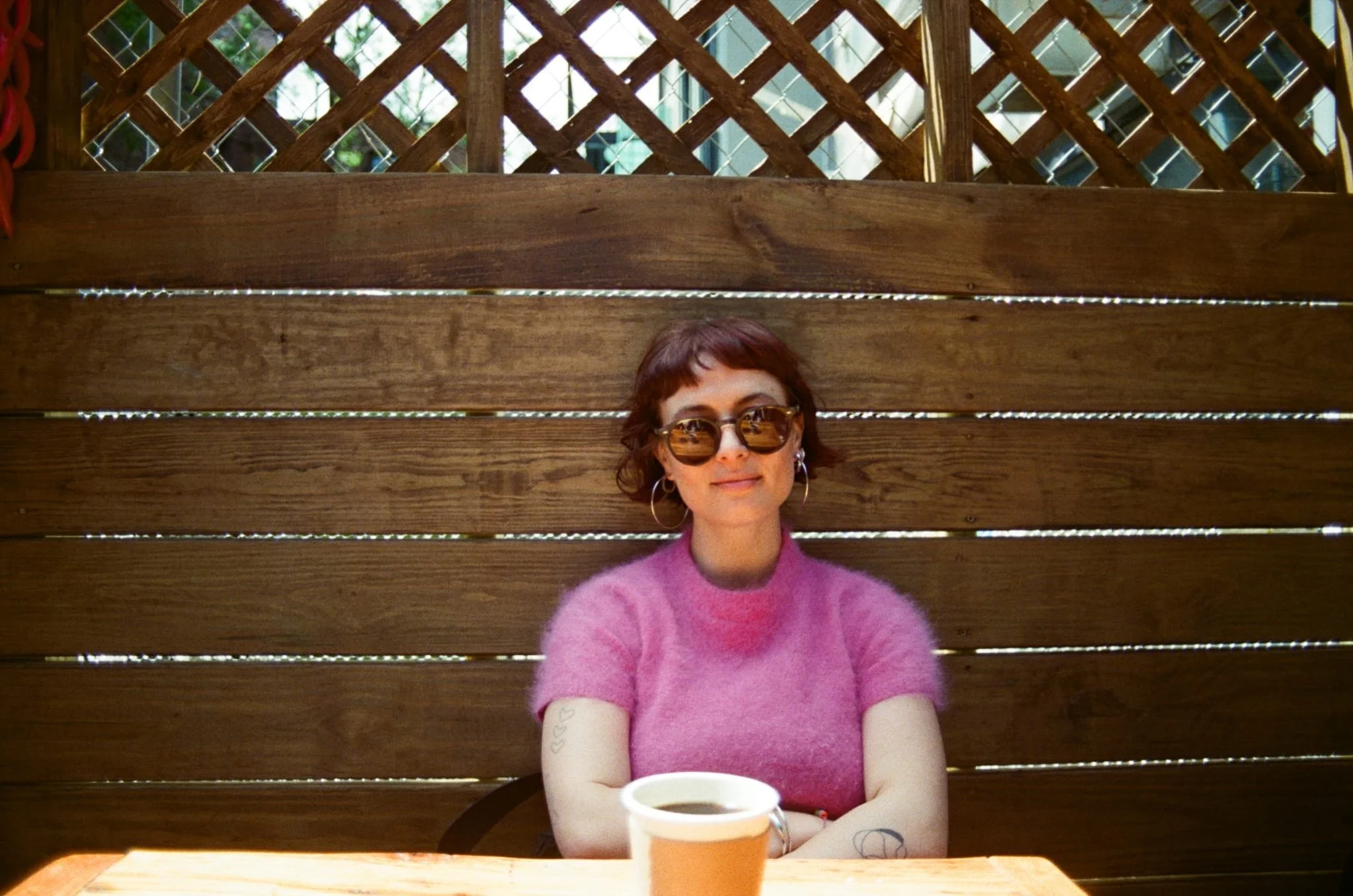 A woman with short dark hair, wearing sunglasses and a pink sweater, is sitting at a wooden table outdoors with a wooden lattice fence behind her and a coffee cup in front of her.