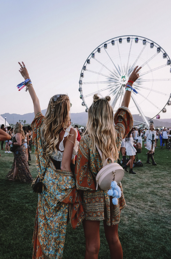 Two women with long, wavy hair standing in front of a Ferris wheel at a festival, with numerous other festival-goers around them, under a clear sky.