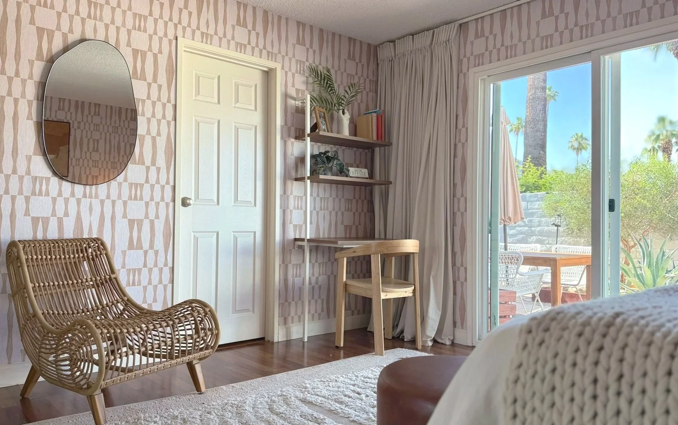 A cozy room with a sliding glass door leading to an outdoor patio with trees and blue sky. The room features a rattan chair, a small wooden table with a chair, a wall-mounted mirror, and a shelf with plants and books. The walls have a geometric patte