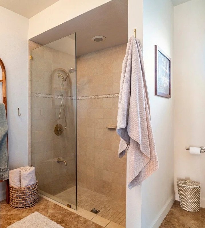 bathroom with a glass-enclosed walk-in shower, beige tiled walls, and a small beige tiled floor. There are towels on a wicker basket on the left, a towel hanging on a hook on the wall, and another towel on the right hanging from a hook. A framed pict