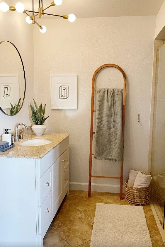Bathroom with a white vanity, potted plant, oval mirror, wall art, wooden towel ladder with gray towel, laundry baskets, and beige rug in Palm Springs vacation rental