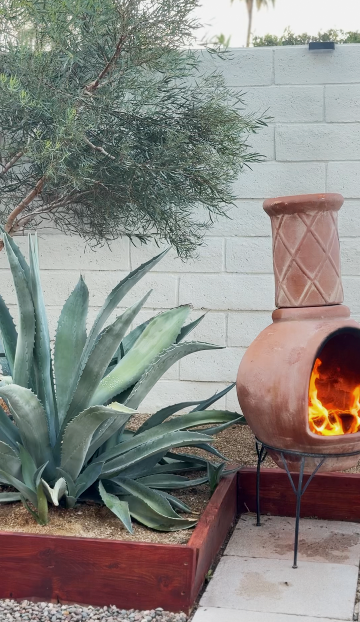 A backyard garden with an agave plant, a small tree, a white brick wall, and a clay chiminea fire pit burning with a flame, all bordered by a red wood frame and concrete pavers.