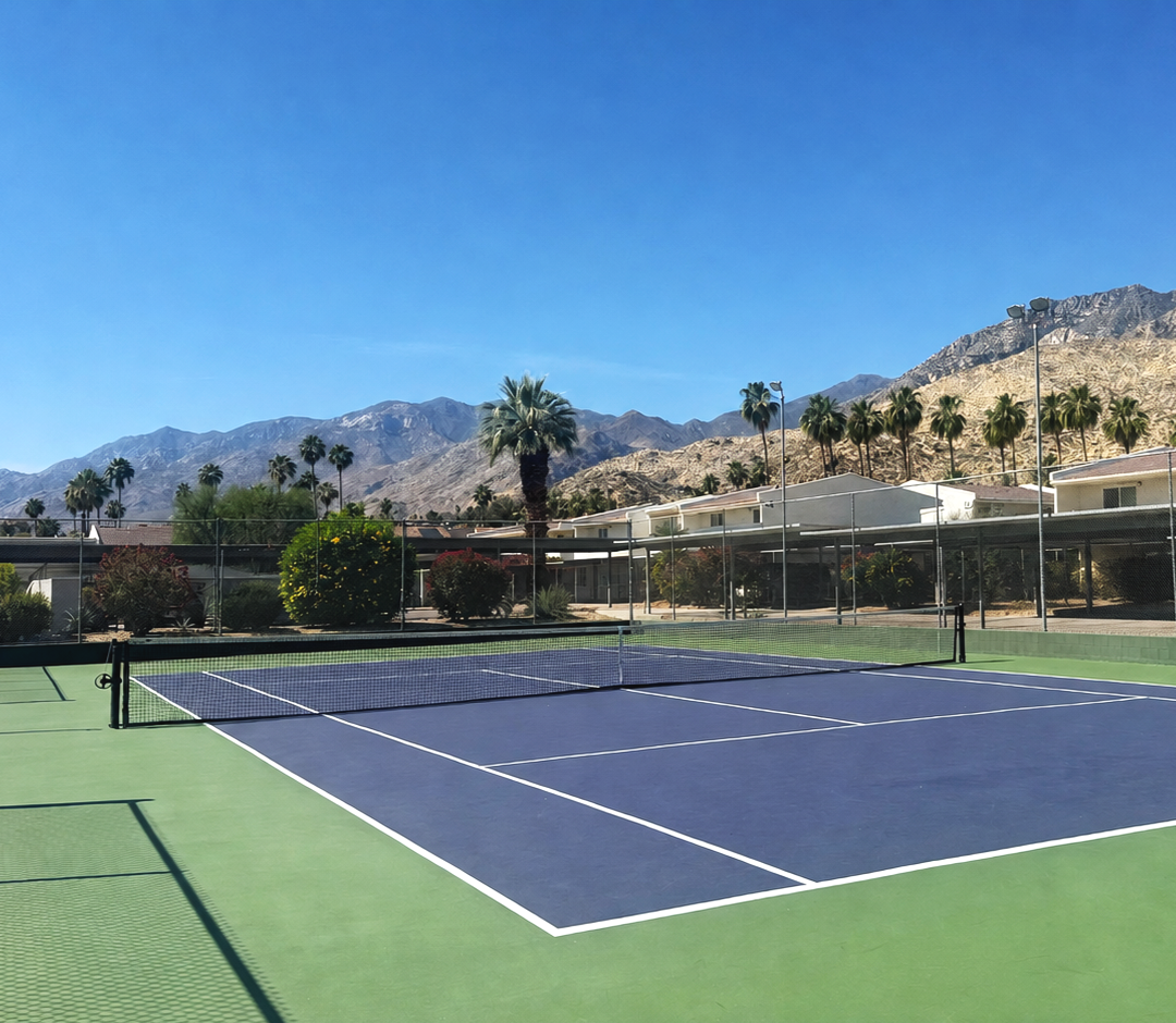 Empty tennis court with green and purple surface, surrounded by palm trees and mountains under a clear blue sky.