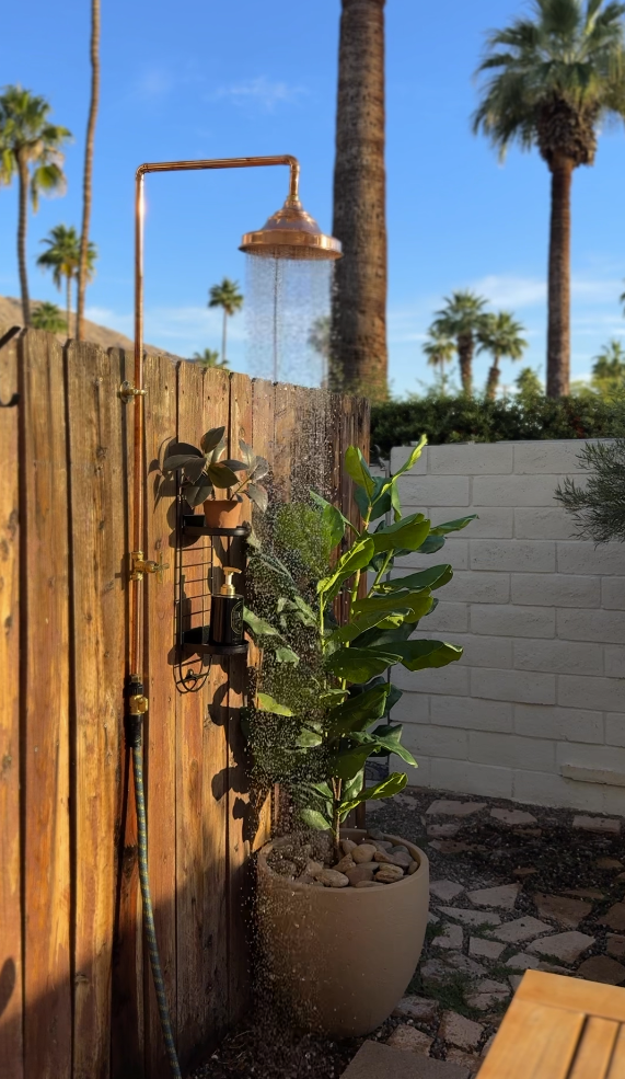 Outdoor shower with water running, wooden fence, potted plant, and palm trees in the background on a sunny day in Palm Springs vacation rental
