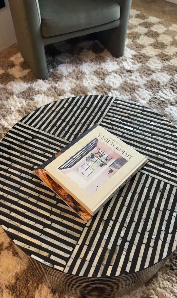 A round black and white striped coffee table with a book titled 'Live Beautiful' on top, in a living room with a beige shaggy rug and a light blue armchair in Palm Springs Vacation Rental