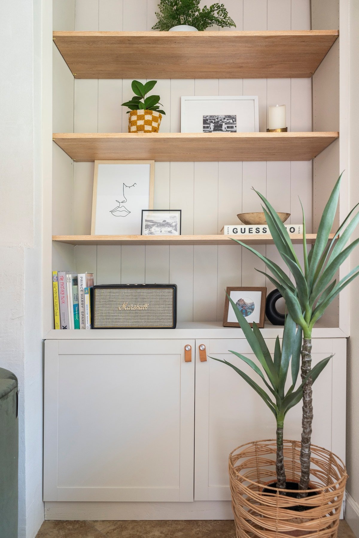 A white cabinet with two closed doors and leather handles, topped with three open wooden shelves holding decorative items, books, and framed photos, with a large potted plant with long green leaves in a wicker basket in front.