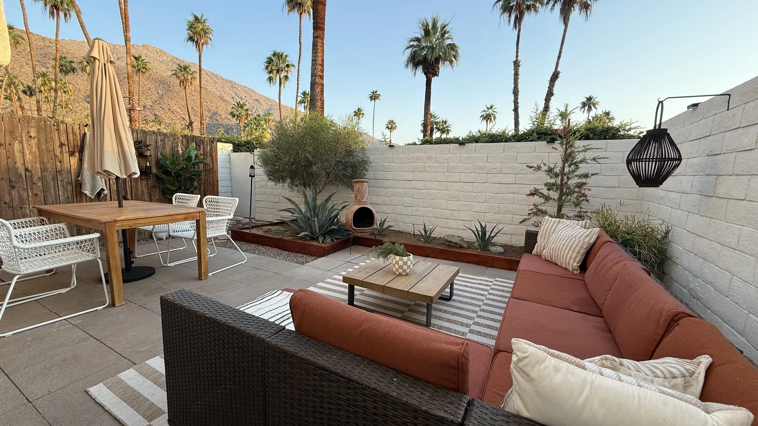 Outdoor patio with a brown wicker sofa with orange cushions, a wooden coffee table, white metal chairs around a wooden table with an umbrella, potted plants, a large agave, a terracotta chiminea, a palm tree, outdoor lighting fixtures, a striped rug,