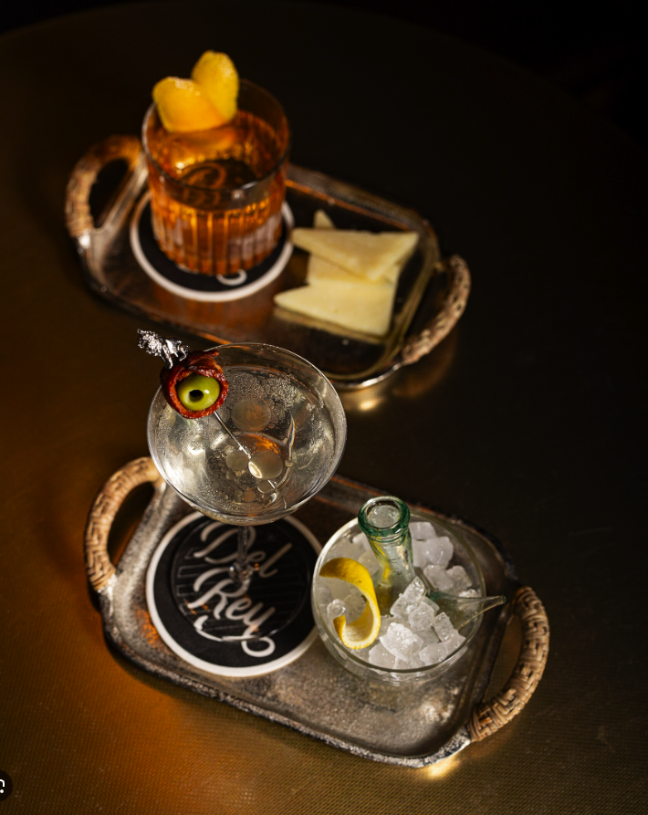 Two beverage trays with drinks and snacks on a dark surface. The tray in the foreground has a cocktail with a cherry and an olive, a bottle of soda in ice, and a lemon wedge. The tray in the background has a glass of whiskey with lemon slices, cheese