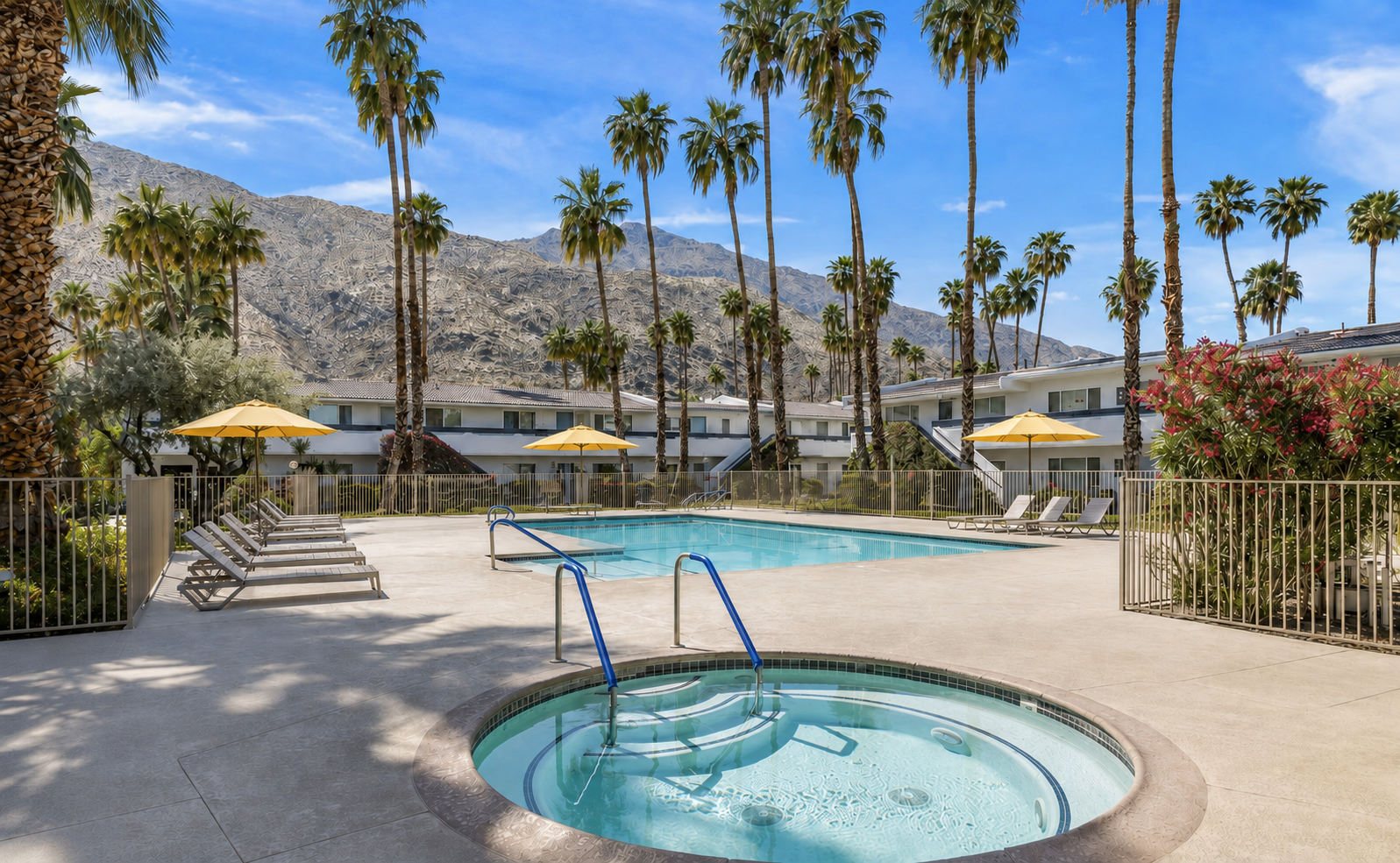 Hotel pool area with lounge chairs, yellow umbrellas, and palm trees, mountains in the background under a blue sky.