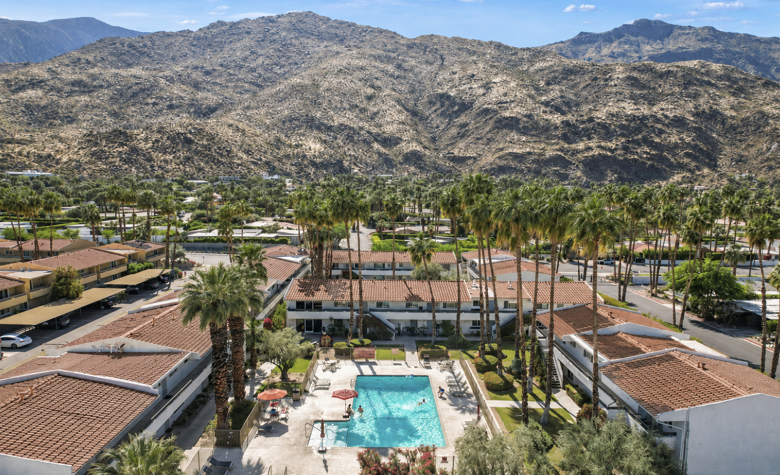 Aerial view of a resort with a swimming pool surrounded by palm trees, white buildings with red tile roofs, and mountainous terrain in the background.