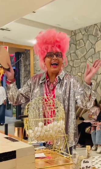 A woman with bright pink afro wig and glasses, wearing a silver sequin jacket, standing behind a yellow raffle drum with white balls, smiling and waving in a lively indoor setting.