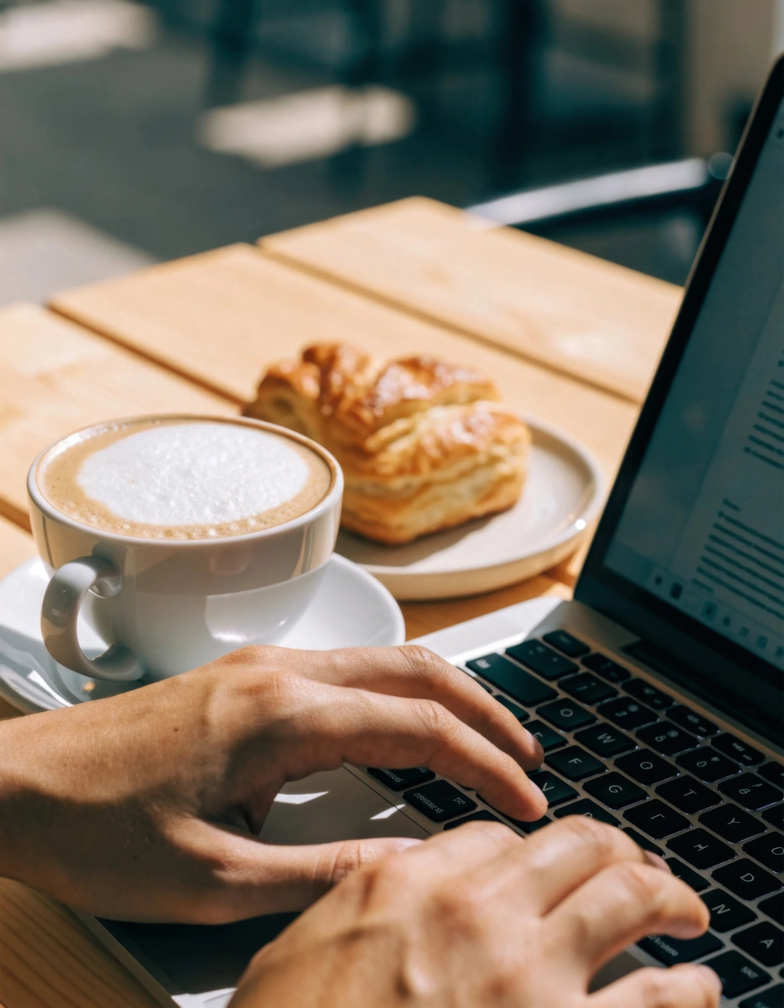 Person typing on a laptop keyboard, with a coffee cup and croissant on a plate nearby, while working at a wooden table, with a laptop screen also visible.