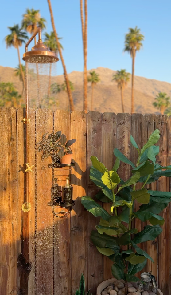 An outdoor shower with a copper rain showerhead mounted on a wooden fence. A potted plant rests on a small black shelf attached to the fence. Sunshine and palm trees are visible in the background, indicating a warm, tropical climate.
