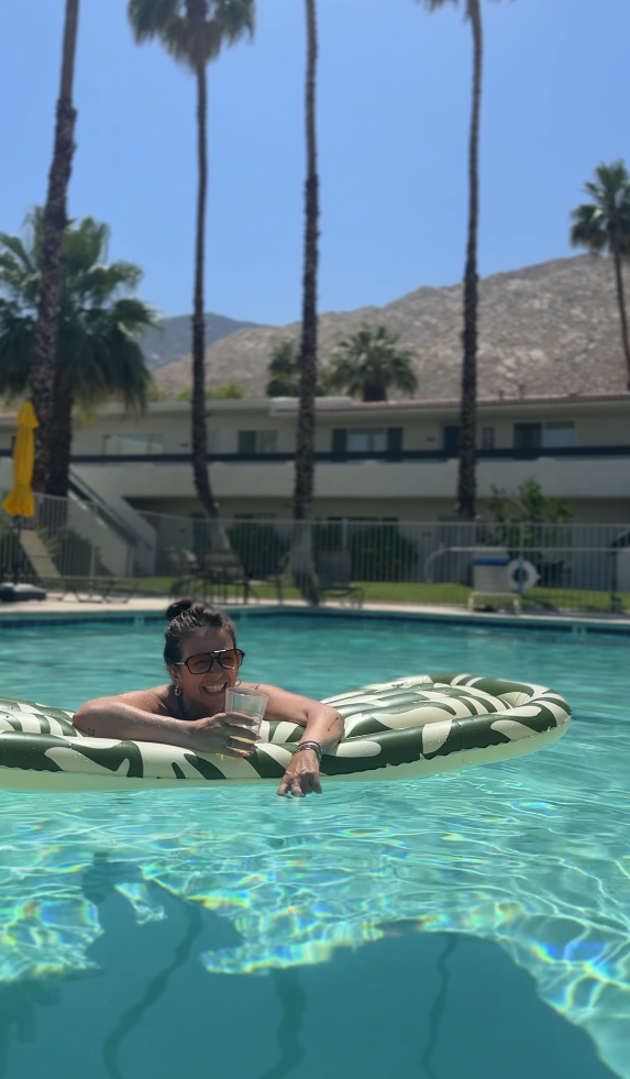 A woman relaxing on a float in a swimming pool, smiling, holding a drink, with palm trees and a clear blue sky in the background.