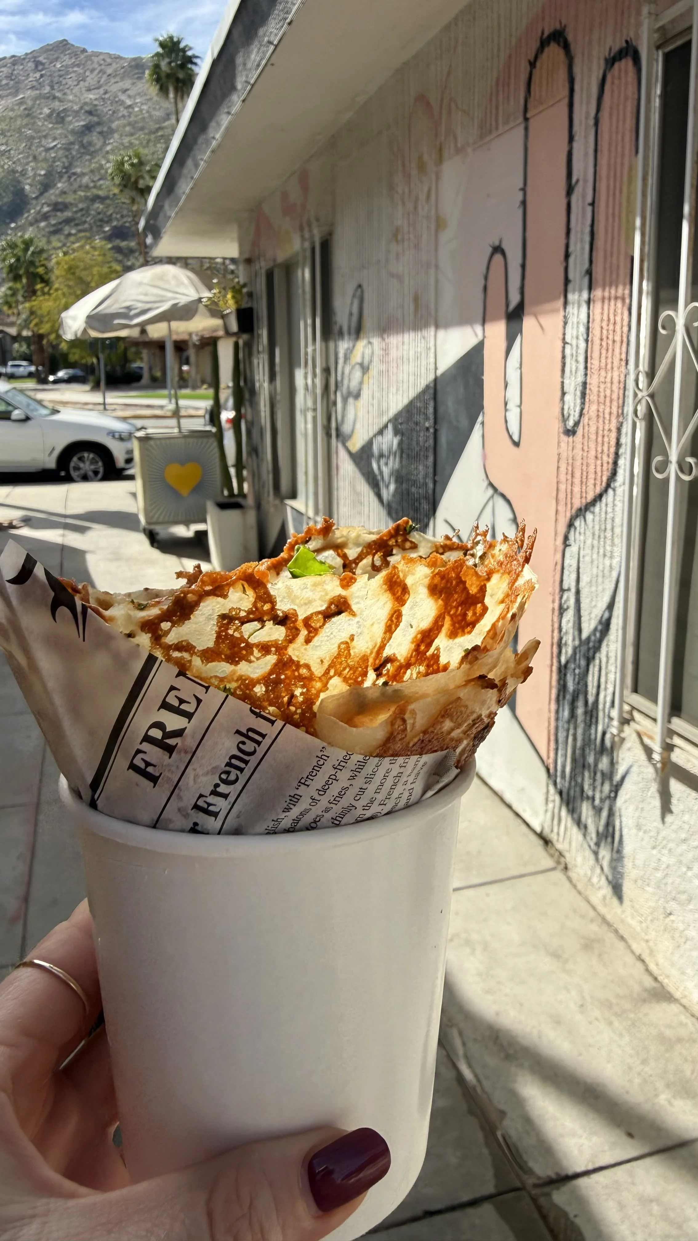 A hand holding a paper-wrapped slice of pizza in a white cup outside a cafe with a mural of abstract faces, hanging plants, and a mountain in the background.