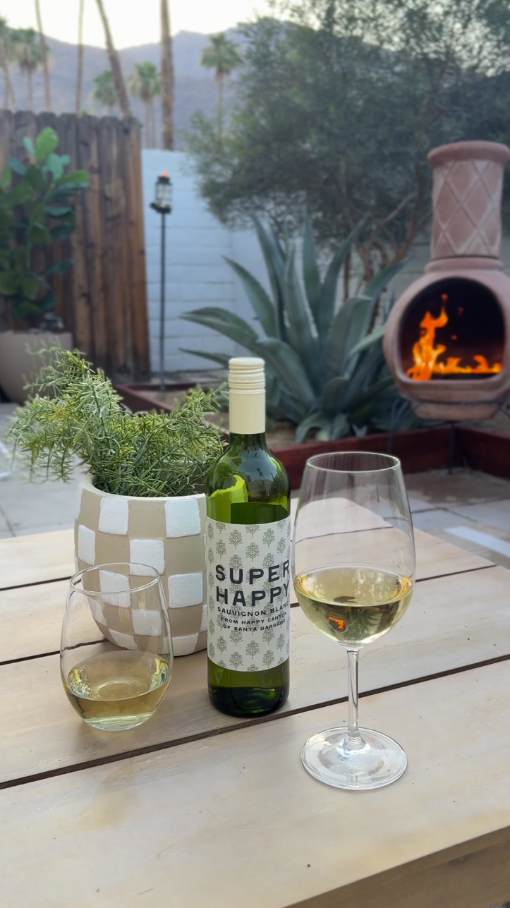 A table with a bottle of white wine labeled 'Super Happy Sauvignon Blanc from Happy Canyon of Santa Barbara,' two glasses of white wine, a potted plant, and an outdoor fireplace with a visible flame, set in a backyard with plants and trees in the background.