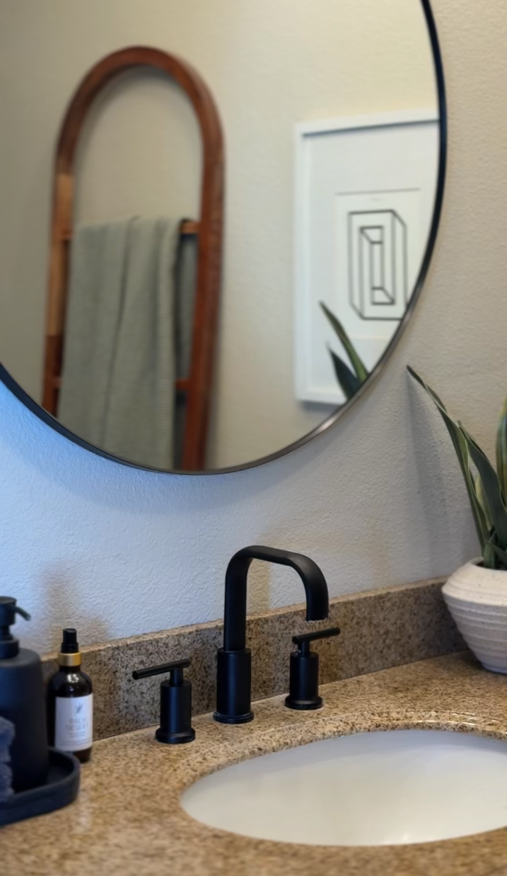 Bathroom sink with black faucet, soap dispenser, potted plant, and a large round mirror reflecting a wooden towel rack and framed artwork on the wall in Palm Springs vacation rental