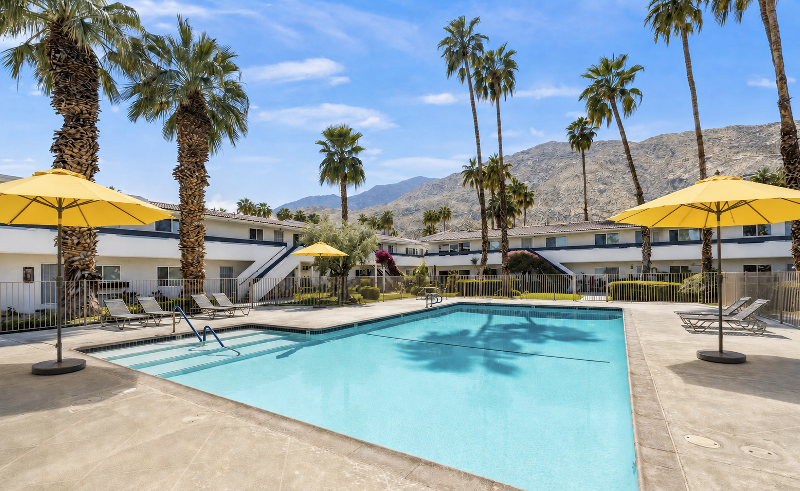 A sunny outdoor pool area with lounge chairs, yellow umbrellas, palm trees, modern buildings, and mountains in the background.