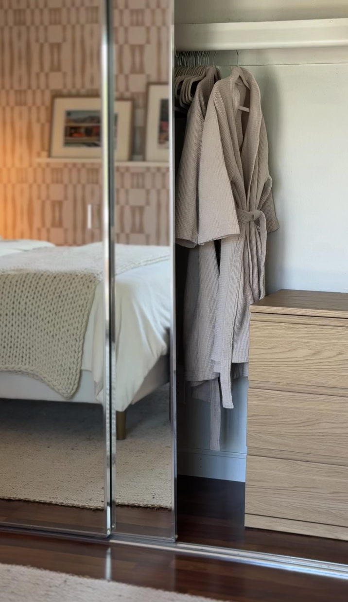A bedroom in Palm Springs vacation rental with a mirrored closet door showing a bed with white linens and a beige blanket, alongside a wooden dresser with beige bathrobes hanging in the closet.