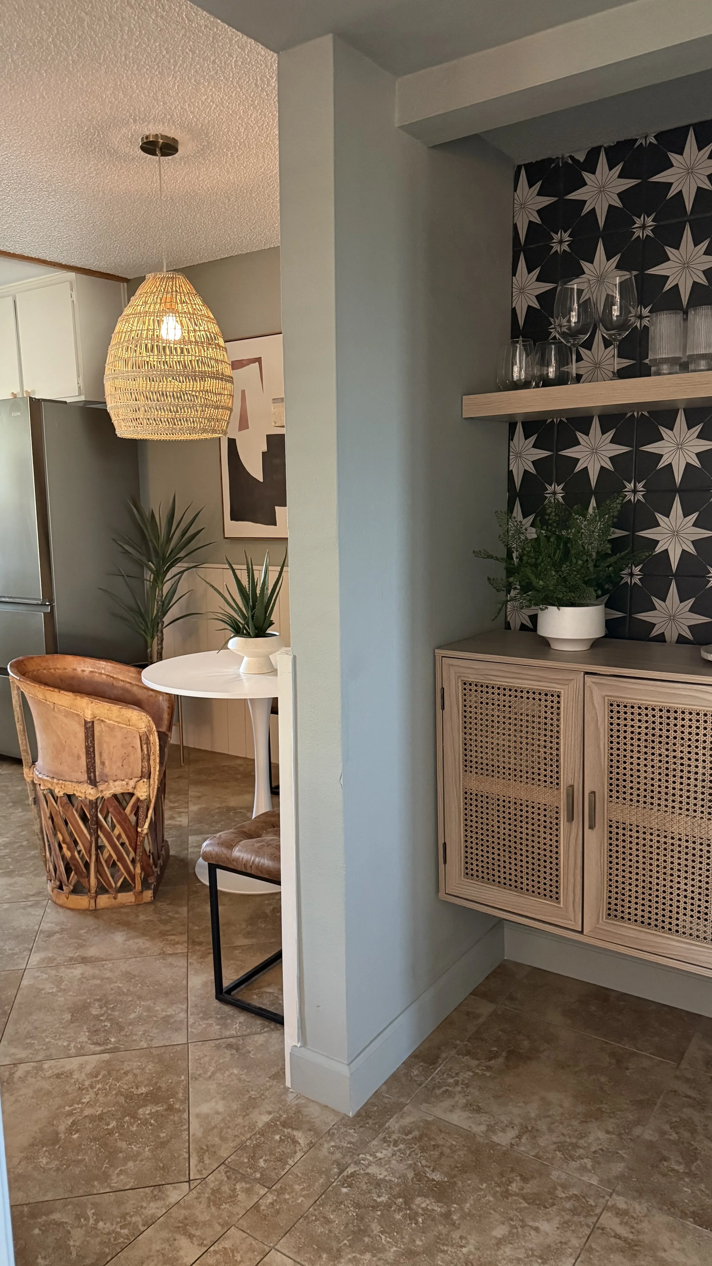 Interior of a modern apartment showing a dining area with a white round table, a vintage style chair, and houseplants. Part of the living room with a hanging woven pendant light and abstract wall art is visible in Palm Springs vacation rental