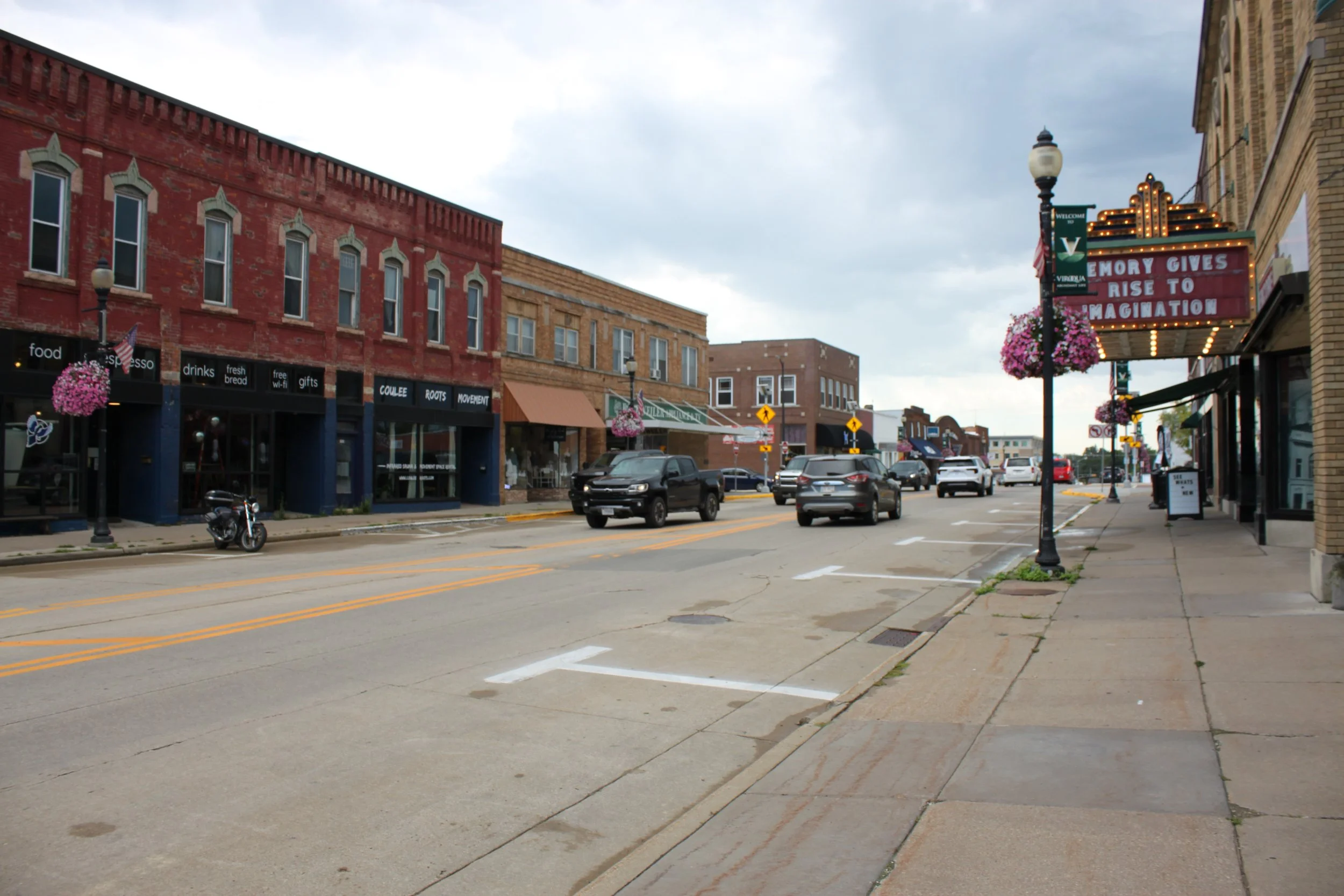 A Viroqua quiet main street with brick and storefront buildings, parked cars, and a motorcycle. The sky is overcast, and decorative flower baskets hang from street lamps. A marquee sign displays the message: 'Memories give rise to imagination.'