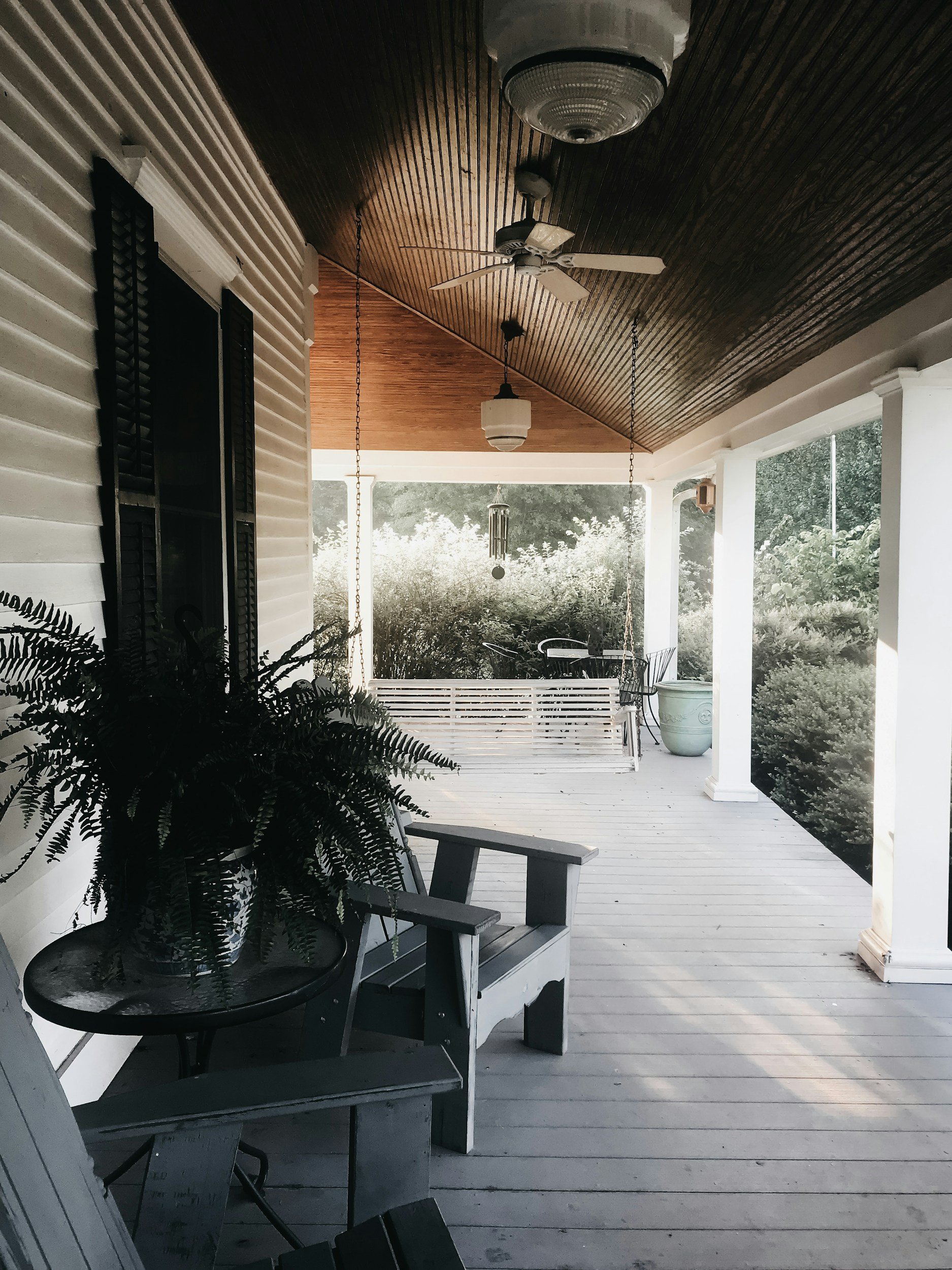 A Viroqua porch with hanging swing, potted plants, ceiling fan, and a ceiling light, overlooking a garden with trees and shrubs.