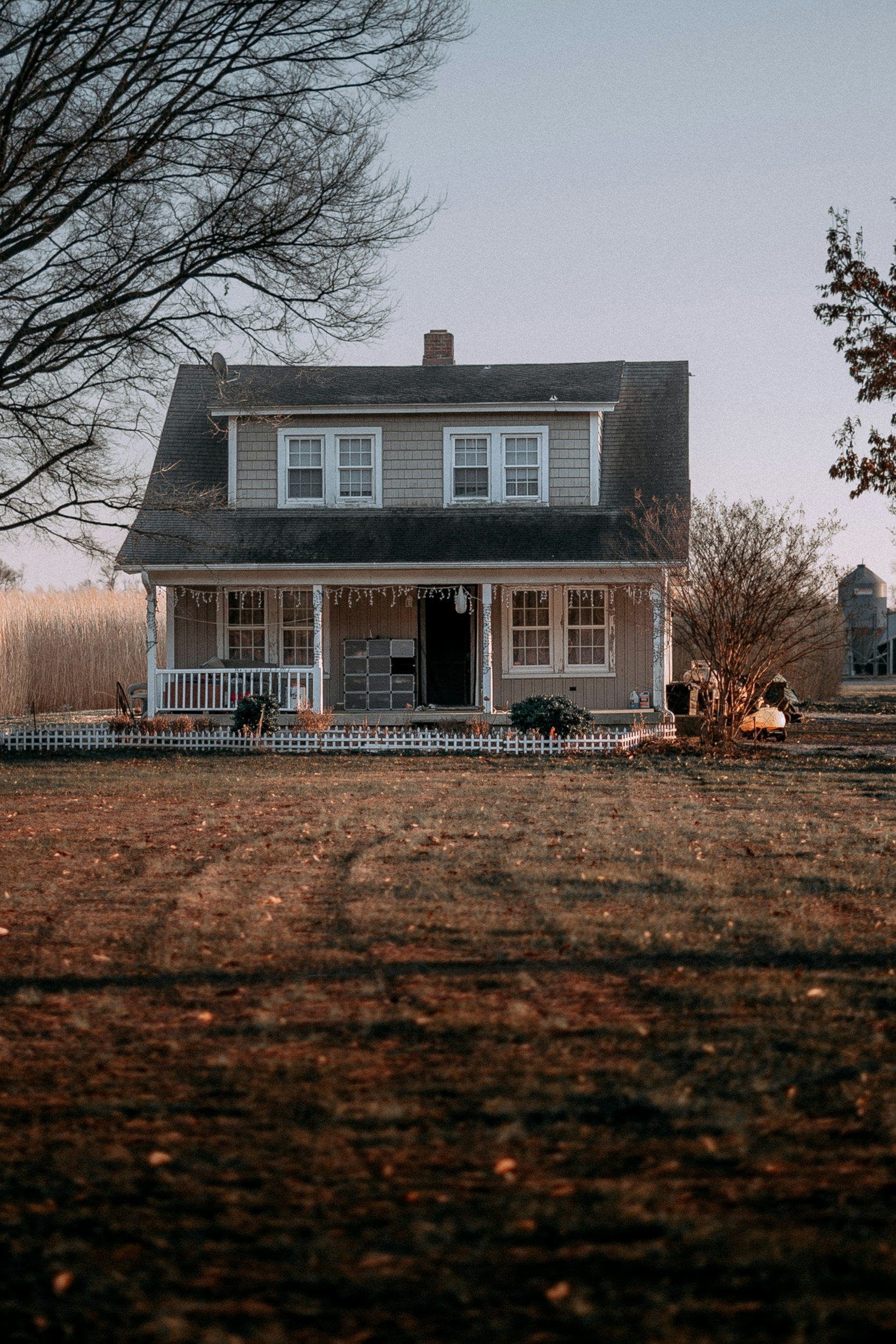 A two-story Vernon County house with gray siding, multiple windows, a front porch with white railing, and a sloped roof, surrounded by trees and a lawn with fallen leaves, during dusk or dawn.