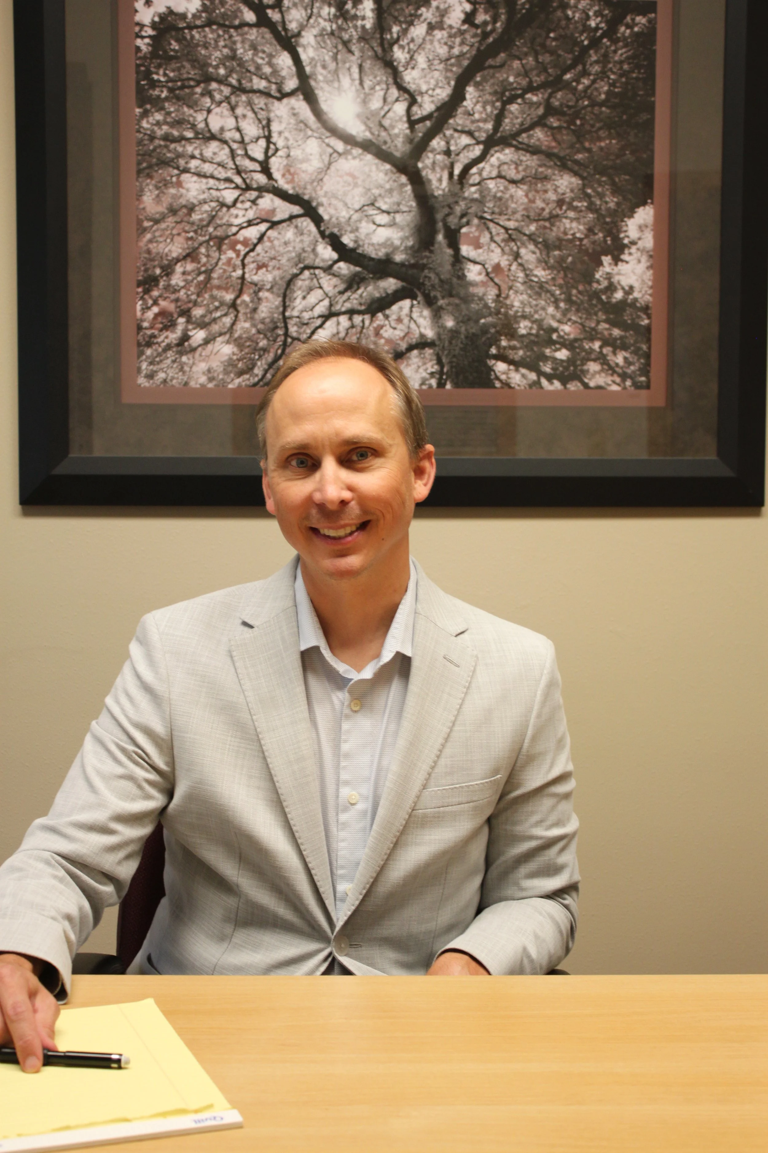 Lawyer Aaron Kane at Viroqua Law Office wearing a light-colored suit sitting at a table with yellow papers, with a framed art photo of a tree in the background.