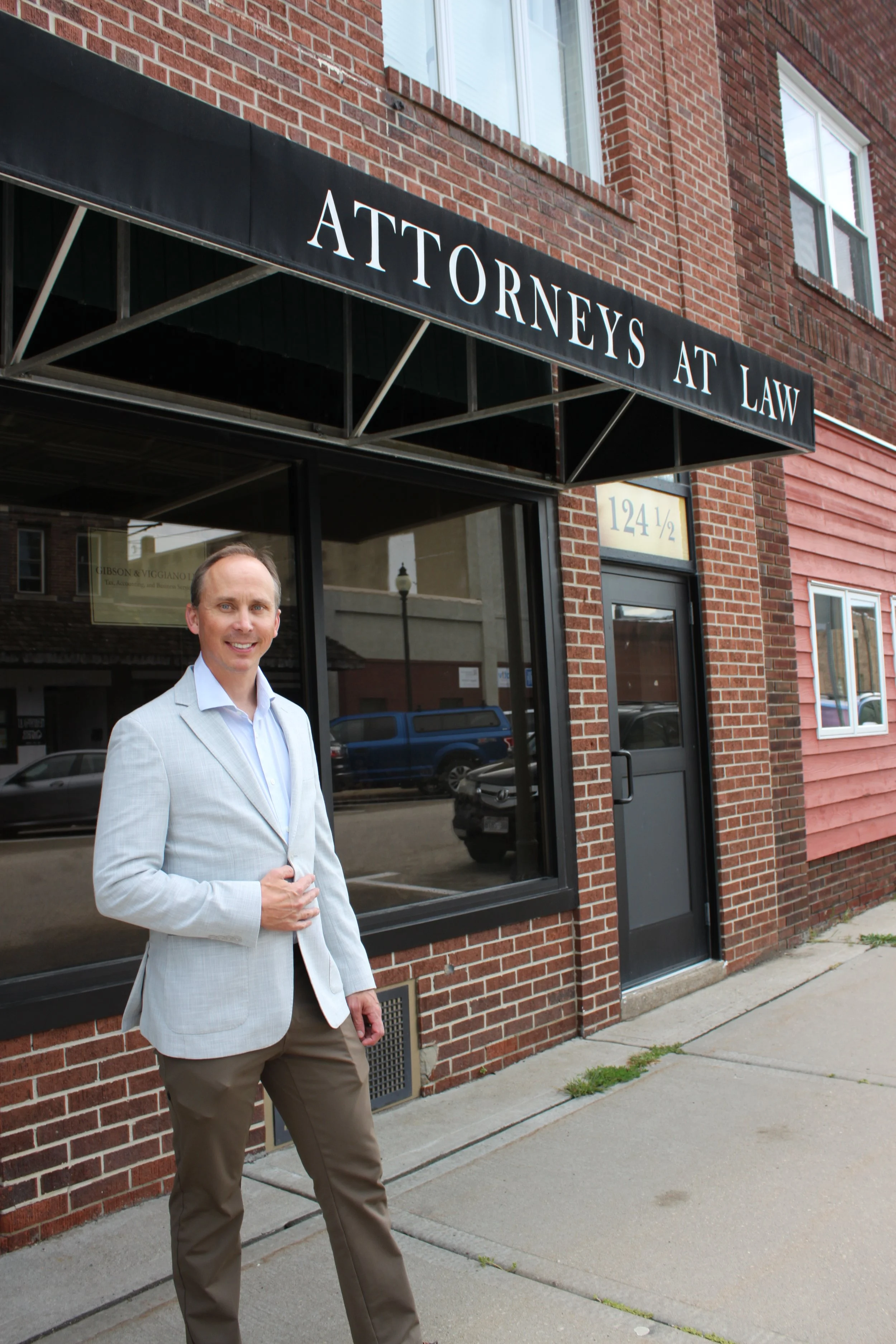 Attorney Aaron Kane in a light blazer and brown pants standing on a sidewalk in front of Land and Legacy Law office - abrick building with a black sign that reads 'ATTORNEYS AT LAW'.
