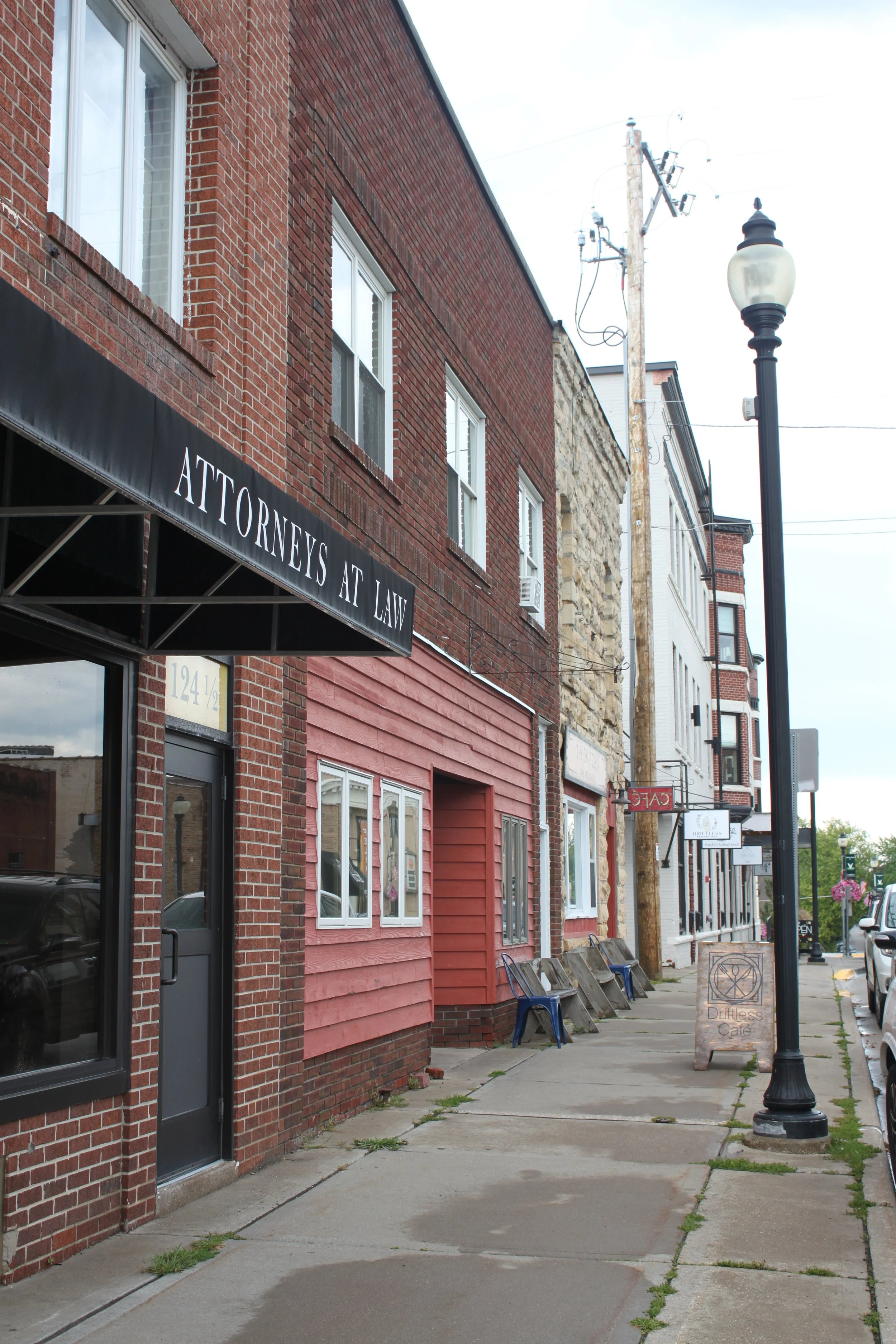 A Viroqua, Wisconsin sidewalk with a sign for Driftless Cafe along a street with brick and wood buildings, a lamp post, and utility poles.