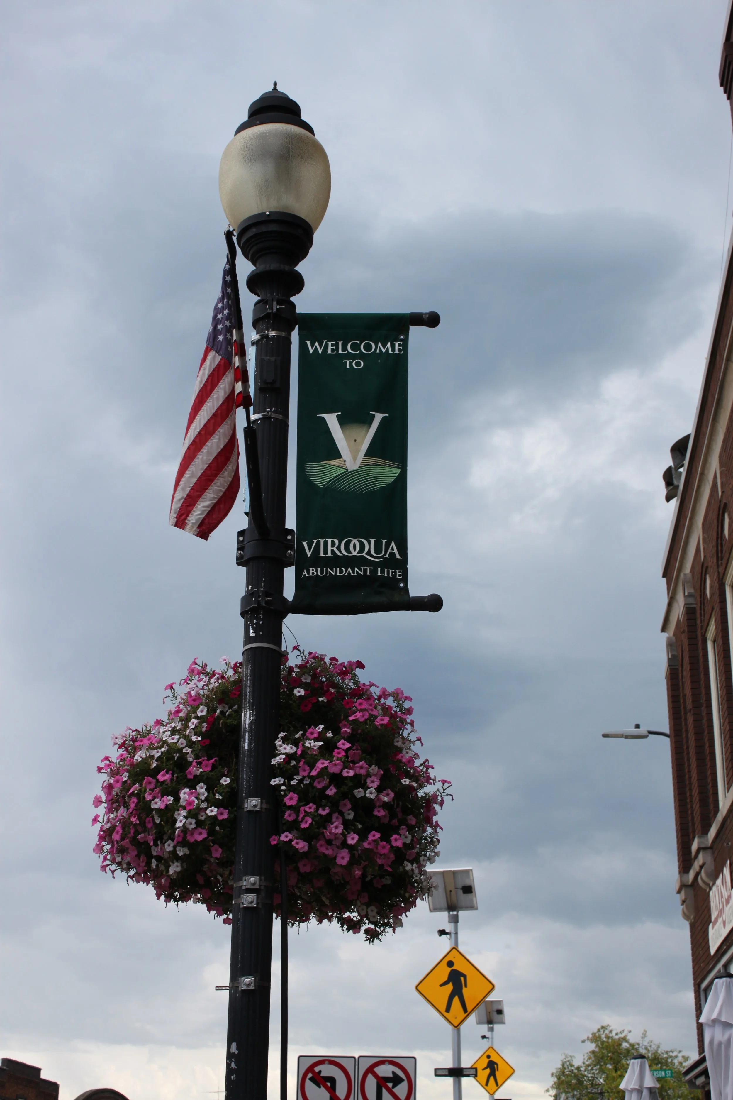 Streetlight with a banner welcoming visitors to Virquua and an American flag, with pink floral basket hanging from the pole, under overcast sky. Pedestrian crossing and no left/right turn signs are visible on the street.