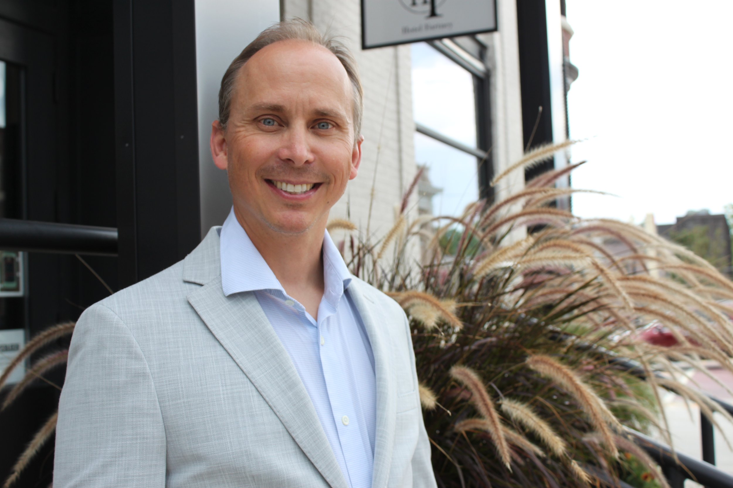 Lawyer, Aaron Kane,  in a light gray suit standing outdoors near tall ornamental grasses, with a Viroqua building and a street sign visible in the background.