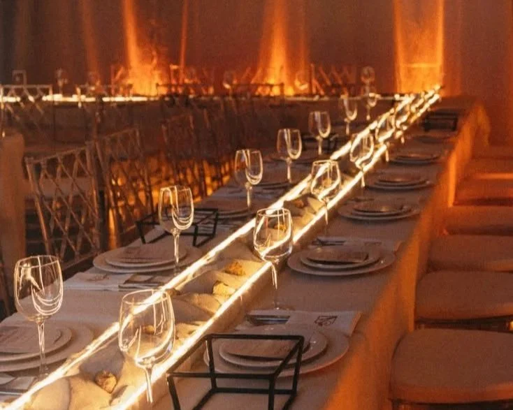 Elegant dining table set with wine glasses, plates, and utensils in a warmly lit room with a long table and chairs.