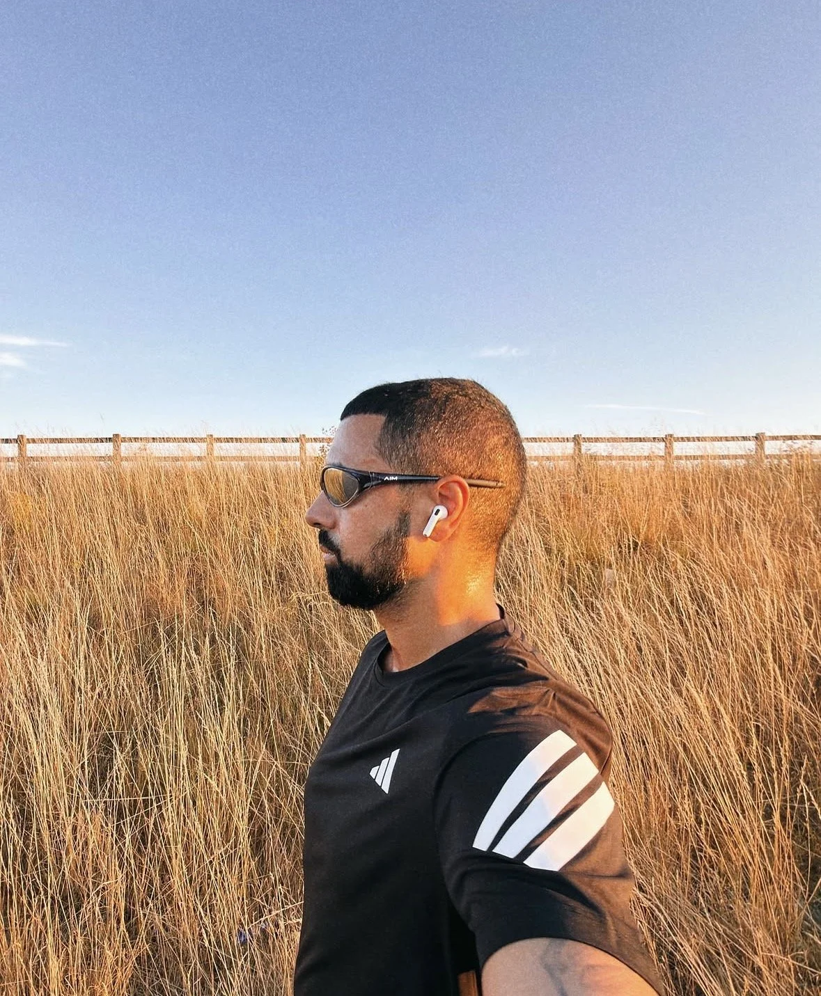 A man with a beard, sunglasses, and wireless earbuds stands in a field of tall golden grass during sunrise or sunset.