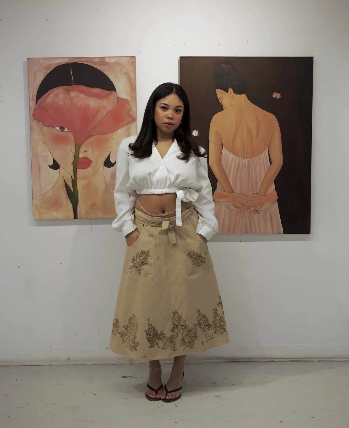 A young woman with dark hair standing in front of two portraits, one of a woman with a flower cover over her face and the other of a woman with her back turned, in an art gallery.