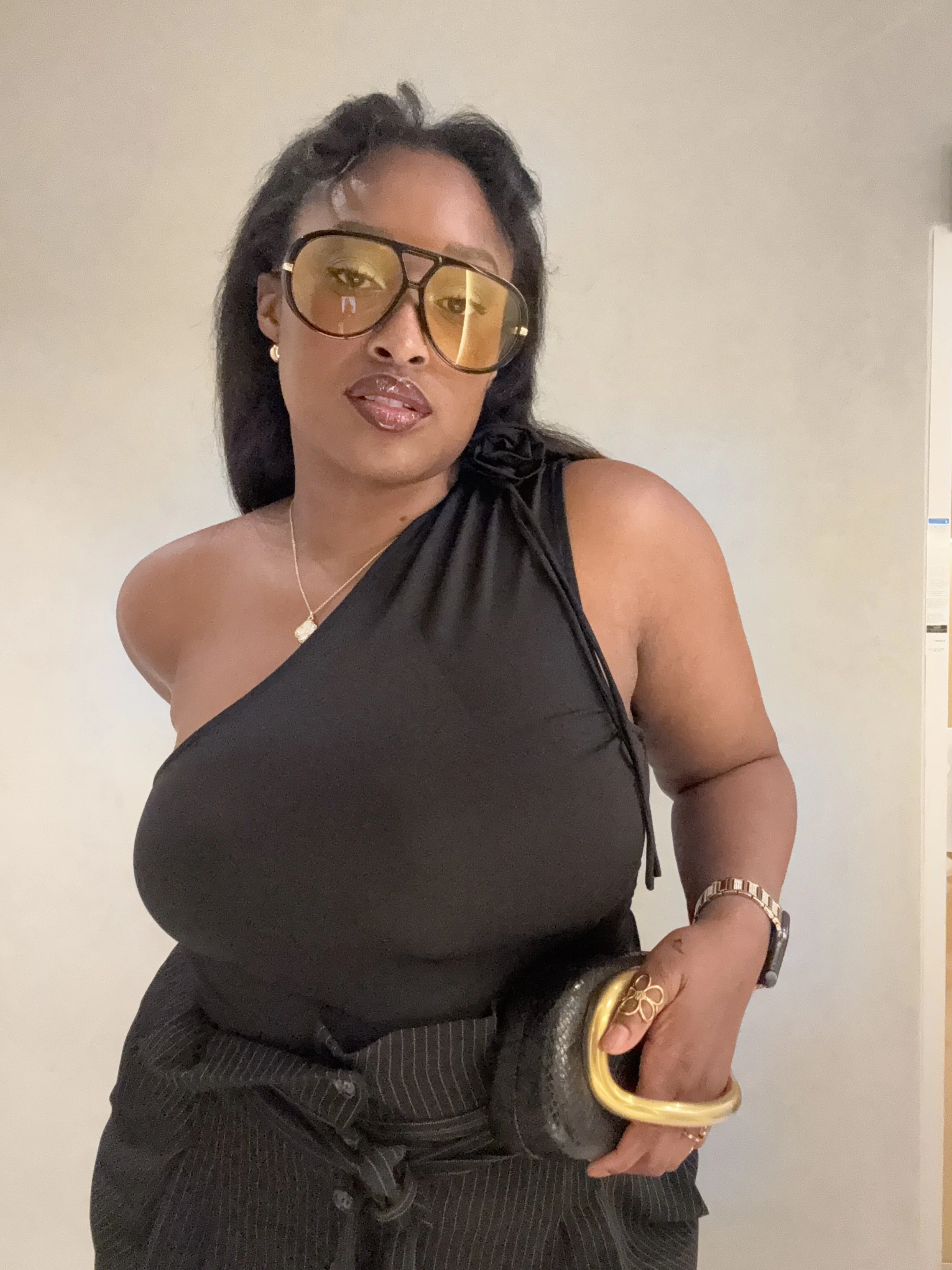 A woman with dark hair wearing large tinted glasses, a black sleeveless top, a gold watch, and jewelry, standing against a plain background.