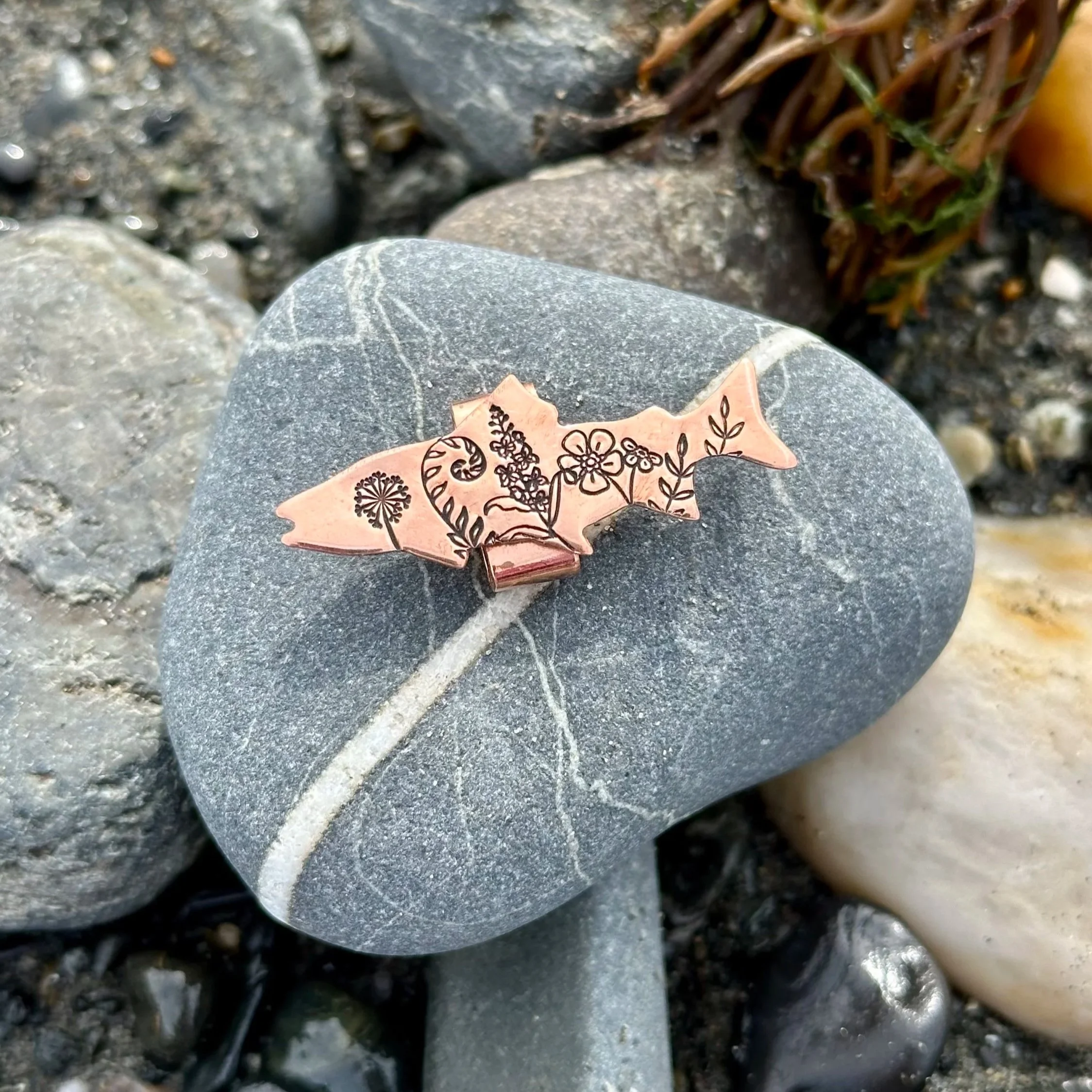 Close-up of grey and black rocks and pebbles with a small decorative copper-colored metal halibut and floral design in the foreground.